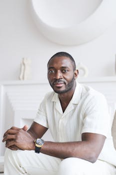 A confident man in white clothing sitting indoors with soft lighting and modern decor.
